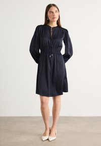 Navy satin dress with a cinched waist, long puff sleeves, and a keyhole neckline detail. Paired with light-colored pointed-toe heels.