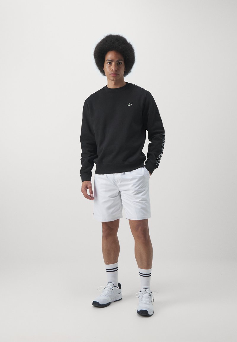 Schwarzes Baumwoll-Sweatshirt mit Rundhalsausschnitt, Logo auf der Brust und Text auf den Ärmeln, kombiniert mit weißen Shorts und Sportschuhen.