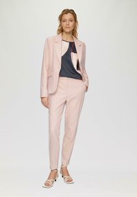 Veste et pantalon rose pâle sur mesure, associés à un haut sombre avec des motifs géométriques. Des sandales à talons blanches complètent l'ensemble.