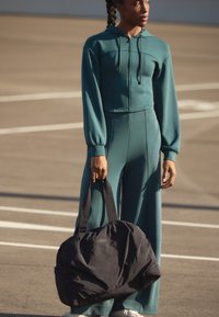 Top cropped à capuche vert canard avec des manches bouffantes, associé à un pantalon large, tenant un grand sac de sport en tissu noir. Texture lisse, design décontracté.