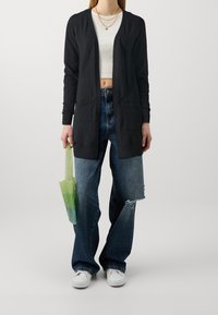 Svart långärmad cardigan, stickat tyg, öppen fram, två framfickor, ihop med slitna vida jeans och en grön gradient tote-väska.