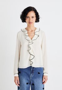 Cream blouse with a deep v-neck, vertical ruffles along the placket, and black trim. Long sleeves with cuffs. Lightweight, slightly sheer fabric.