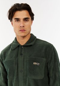 Mladic s tamnom kosom nosi tamnozeleni duksericu na dugmad s džepovima na prsima i oznakom "PROTEST" na lijevom džepu.