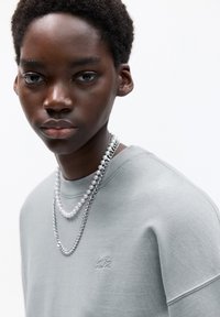 Sivá sweatshirt z teksturirane tkanine, ki ima majhen izvezeni logotip. Plastični ovratniki iz biserov in kovinske verige dodajajo kontrast.