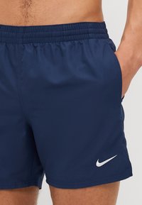 Sötétkék úszónadrág elasztikus derékkal, oldalsó zsebekkel és egy fehér Nike logóval az alsó részen. Simaság és könnyű anyag.