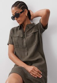 Kurzärmliges, olivgrünes Leinenhemdkleid mit zwei Brusttaschen, Knopfleiste, lässiger Passform, getragen mit übergroßen schwarzen Sonnenbrillen.