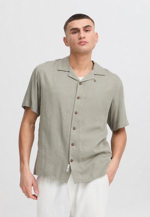 Homme portant une chemise grise claire à manches courtes boutonnée et un pantalon blanc, debout devant un fond clair uni.