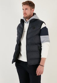 Mjuk black ärmlös väst med dragkedja, prydd med ett chevronmönster och en rund logotyp på bröstet, bärs över en hoodie.