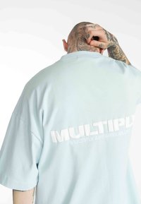 Světle modré oversized tričko vyrobené z měkké tkaniny, s bílým embosovaným nápisem "MULTIPLY APPAREL CLUB" na zadní straně. Krátké rukávy.