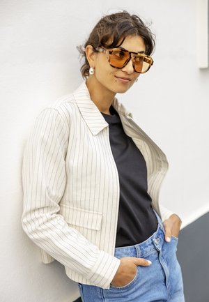 Žena s krátkymi kučeravými vlasmi, ktorá nosí veľké okuliare so vzorom korytnačky, biele saka s tenkými pruhmi, čiernu košeľu a modré džínsy, ruky v vreckách.