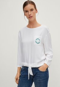 Camicia bianca a maniche lunghe con scollatura rotonda, cintura regolabile in vita e tasca con testo ricamato blu. Abbinata a jeans blu.