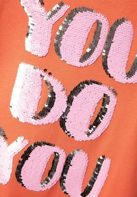 Tela naranja con grandes letras de lentejuelas que dicen "TÚ HACES LO QUE QUIERES". Las lentejuelas rosas y negras crean un contraste brillante.