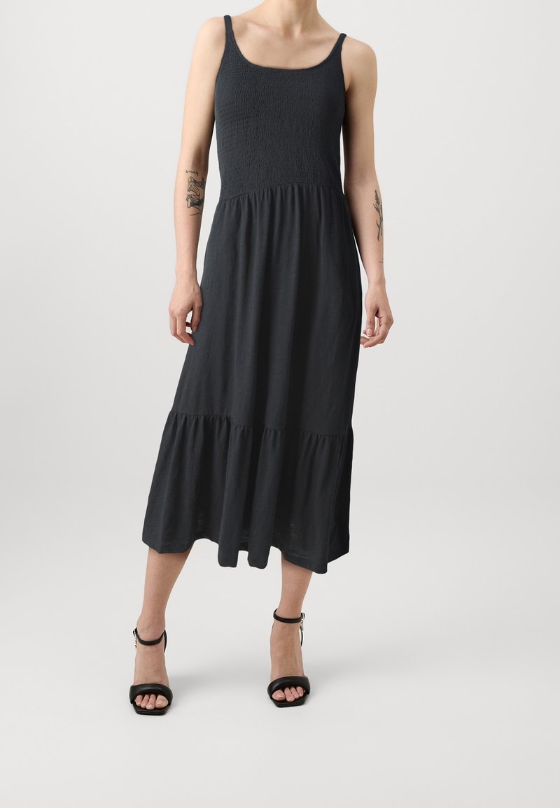 Personne portant une robe midi noire sans manches avec un corsage smocké et un ourlet volanté, associée à des sandales à talons noires à bride cheville.