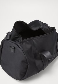 Bolso de deporte negro hecho de tejido texturizado, con una parte superior abierta espaciosa, correas planas y un pequeño detalle de hardware en el lateral.