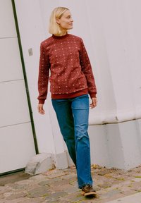 Sudadera roja con un patrón, de cuello alto, combinada con jeans rectos azules y zapatos marrones, caminando sobre adoquines.