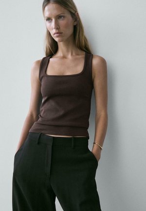 Giovane donna con capelli lunghi che indossa un top marrone senza maniche con scollo quadrato e pantaloni neri, in piedi davanti a uno sfondo chiaro e uniforme.