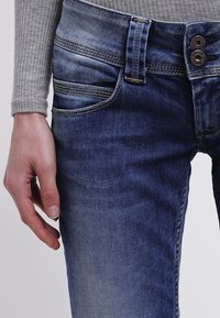 Närbild av en persons vänstra hand som vilar bredvid mörkblå urtvättade jeans med dubbelknappsstängning och kontrastsöm.