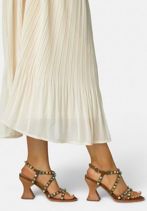 Sandalias de tacón marrones con gemas multicolores, usadas con una falda midi plisada color crema, mostrando los pies y las pantorrillas inferiores.