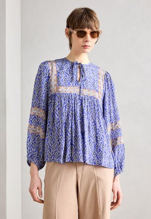 Blauwe blouse met een patroon, een sleutelgat-halslijn, plooidetail en lange mouwen; gecombineerd met lichtbeige wijde broek.