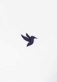 Tejido blanco con un pequeño logo de colibrí bordado en azul oscuro en el lado izquierdo, mostrando detalles en las alas y el cuerpo.
