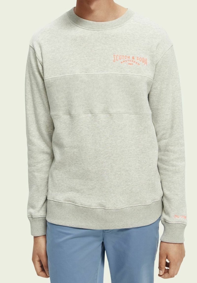 Sweat-shirt gris clair à col rond avec le logo « Scotch & Soda Amsterdam 1985 » en orange sur la poitrine, porté avec un pantalon bleu clair.