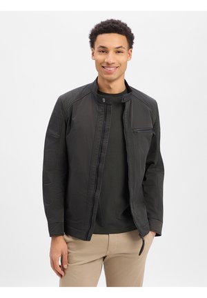 Lächelnder junger Mann, der eine schwarze Jacke mit Reißverschluss über einem dunklen Hemd und beigen Hosen trägt, stehend mit teilweise in den Taschen befindlichen Händen.