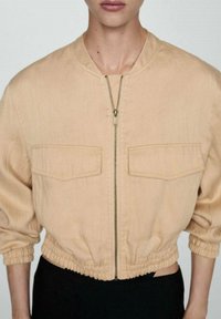 Jasnobeżowa krótka kurtka zapinana na zamek z elastycznymi mankietami i dwoma kieszeniami na piersi; wykonana z gładkiej tkaniny o subtelnej fakturze.