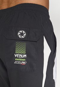 Sorte træningsshorts med en baglomme med flap, trykte logoer i hvid og grøn, samt hvide sidelåse. Tekstur detajle.