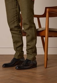 Zwarte leren dresschoenen met een gladde textuur en een spitse neus, gecombineerd met donkergroene getextureerde broek, tegen een achtergrond van een houten stoel.