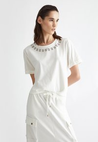 T-shirt bianco a maniche corte con scollatura rotonda e un motivo decorativo di piccoli accenti metallici. Tessuto morbido e vestibilità comoda.