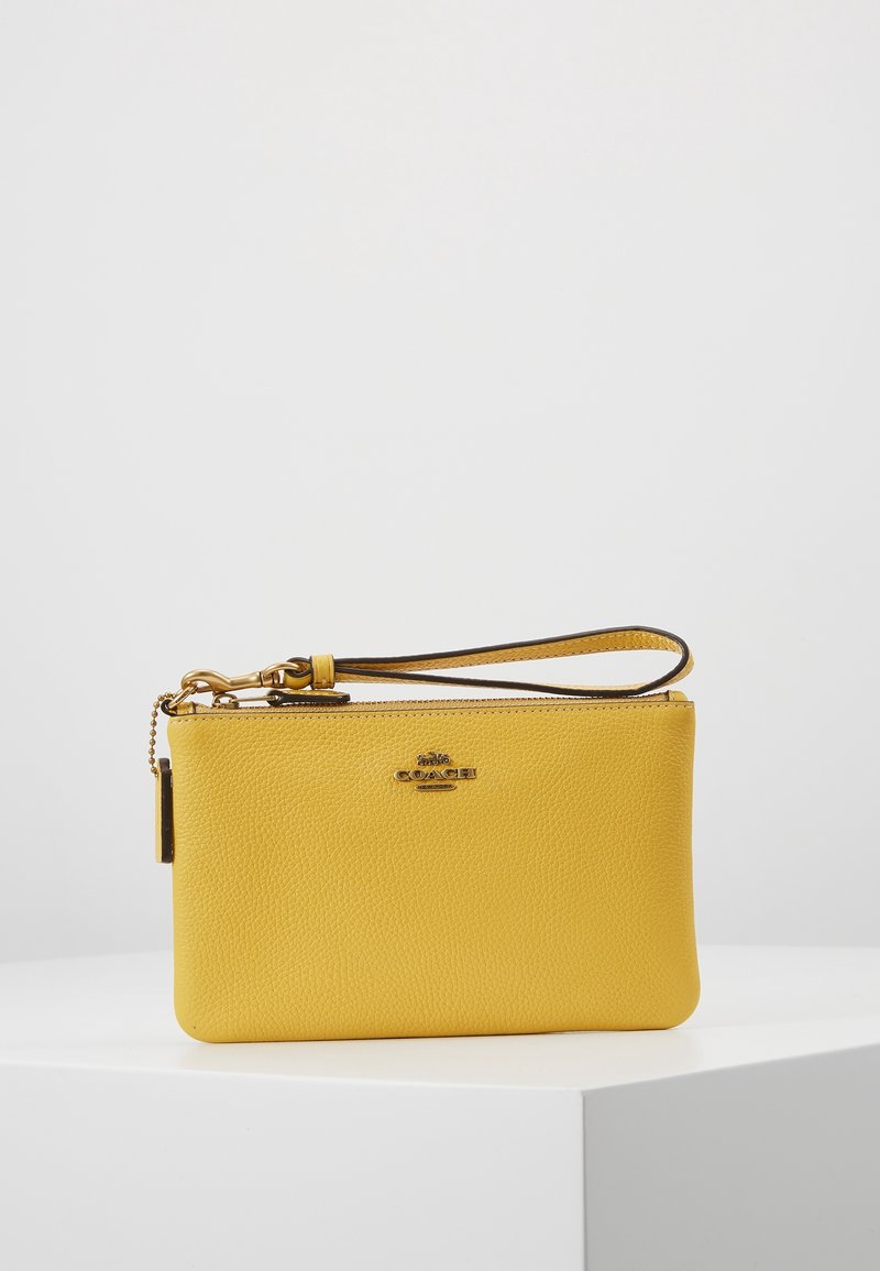 Pochette bracelet en cuir texturé jaune avec fermeture éclair dorée, logo Coach doré et bracelet assorti sur une surface blanche avec un fond gris.