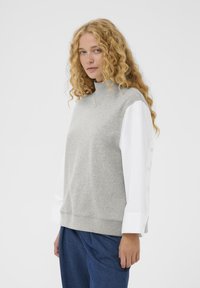 Sweat-shirt gris à col montant, manches de chemise blanches avec détails de boutons. Texture douce, coupe décontractée, associé à un pantalon bleu.