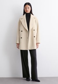 Dobbeltknappet, cremefarvet frakke lavet af tekstureret stof, med store brune knapper og klapkrave, båret over en sort turtleneck.
