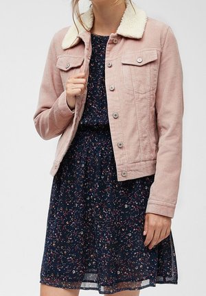 Femme portant une veste en velours côtelé rose avec un col en polaire blanche, par-dessus une robe courte à fleurs de couleur bleu foncé.