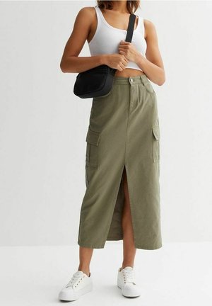 Kvinde iført en hvid cropped tank top, olivengrøn cargo midi nederdel med frontslids, hvide sneakers og bærer en sort crossbody taske.