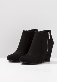 Czarne zamszowe botki na koturnie sięgające do kostek, z bocznymi zamkami błyskawicznymi, umieszczone na białej powierzchni na tle jasno szarego koloru.