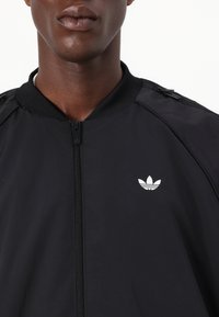 Melns jaku ar rāvējslēdzēju un ribotu apkakli, ar baltu Adidas logotipu kreisajā krūtī un rāvējslēdzējiem uz pleciem.