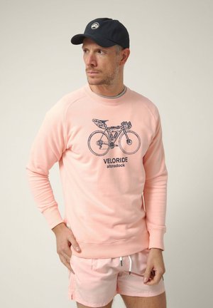 Hombre con sudadera rosa claro con gráfico de bicicleta y pantalones cortos a juego, llevando gorra de béisbol oscura, mirando hacia un lado con fondo liso.