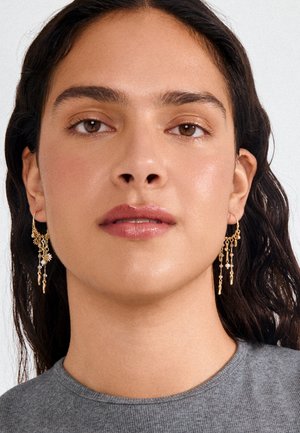 Žena s tamnom valovitom kosom nosi viseće zlatne naušnice s malim privjescima i sivu rebrastu košulju, okrenuta prema naprijed na jednobojnoj pozadini.