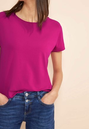 Kobieta ubrana w jaskraworóżowy t-shirt z krótkim rękawem, włożony do niebieskich dżinsów, z rękami w kieszeniach na jasnym tle.