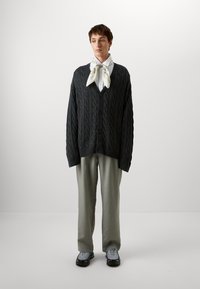 Dark gray cable-knit cardigan with long sleeves, button front, worn over a light shirt and white patterned scarf; paired with gray trousers and sneakers.