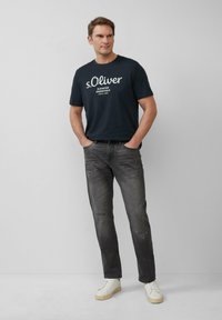 Granatowa koszulka z białym logo, zestawiona z przecieranymi szarymi dżinsami z wy\u017cyszeniami oraz białymi trampkami, zaprezentowana na neutralnym tle.