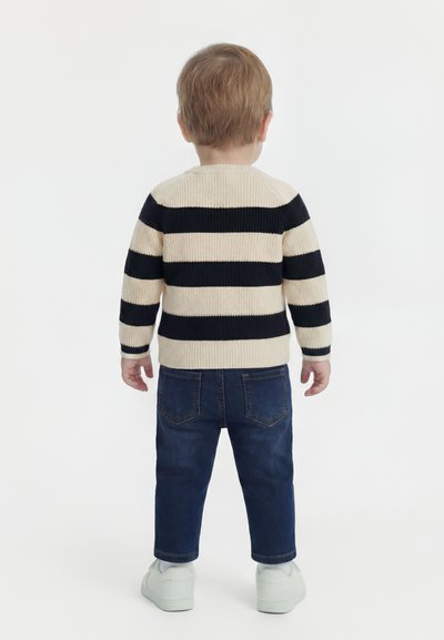 Μικρό παιδί που φοράει πουλόβερ με κρεμ και μαύρες ρίγες, σκούρο μπλε τζιν και λευκά αθλητικά παπούτσια, στέκεται με την πλάτη προς τα εμπρός πάνω σε λευκό φόντο.