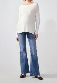 Top blanco de punto con mangas largas y aberturas laterales, combinado con jeans de campana azules. Unos zapatos planos negros completan el atuendo, mostrando un estilo informal.