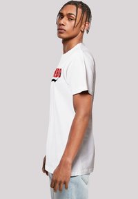 White cotton t-shirt with round neck, featuring red text and a black graphic design. Worn with light blue denim jeans, side profile displayed.
