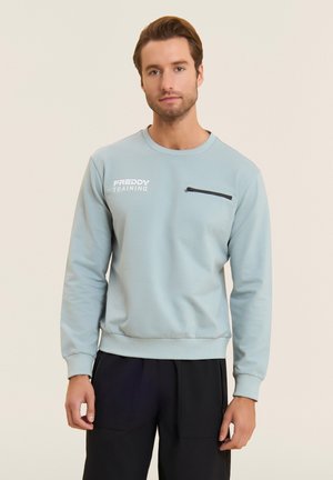 Mies pukeutunut vaaleansiniseen collegepaitaan, jossa lukee "FREDDY TRAINING" ja musta vetoketjullinen rintatasku, yhdistetty mustiin housuihin, kasvot eteenpäin.