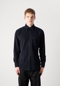 Camisa de pana azul oscuro con botones, con un bolsillo de parche, mangas largas y tejido texturizado, combinada con pantalones negros.