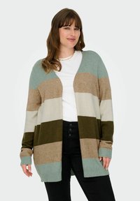 Svītrots džemperis ar mīkstu audumu zaļā, beige un krēmkrāsā, ar brīvu piegriezumu, atvērtu priekšpusi un ribotām aprocēm. Nēsāts virs baltas blūzes.