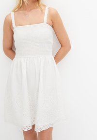 Vit, smocksyd klänning med fyrkantig halsringning, tunna axelremmar och volangkanter. Kjolen har utskurna spetsdetaljer vid fållen.