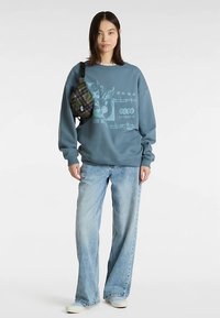 Blauwe trui met grafische print aan de voorkant, gecombineerd met lichtblauwe jeans met wijde pijpen. Accessoires zijn een geruite heuptas en lichtkleurig schoeisel.